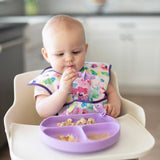 Baby in high chair with colorful bib holds Bumkins Silicone Chewtensils®: Lavender, enjoying food from a sectioned plate. Kitchen behind.