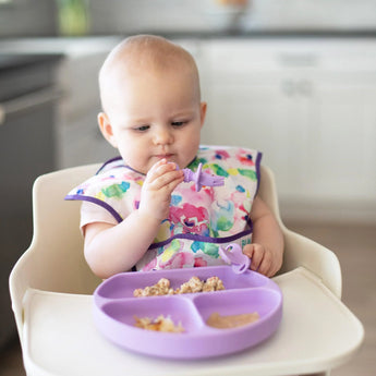 A baby in a colorful bib uses Bumkins Silicone Chewtensils®: Lavender in a high chair with a purple divided plate exploring weaning.