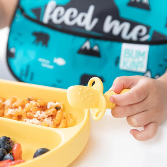 A child with a feed me bib holds a yellow Bumkins Silicone Chewtensils®: Pineapple by a plate of food.