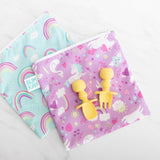 Two colorful unicorn and rainbow bags on a white surface; Bumkins Silicone Chewtensils®: Pineapple fork and spoon rest atop one bag.