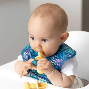 A baby enjoys Bumkins Silicone Chewtensils®: Pineapple, using a blue bib and yellow spoon for baby-led weaning.