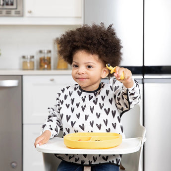 Curly-haired child in a high chair smiles with a spoon, heart bib, and Bumkins Silicone Chewtensils®: Pineapple food-safe plate.