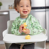 A happy toddler in a high chair, wearing a green bib, enjoys baby-led weaning with Bumkins Silicone Chewtensils®: Pineapple in the kitchen.