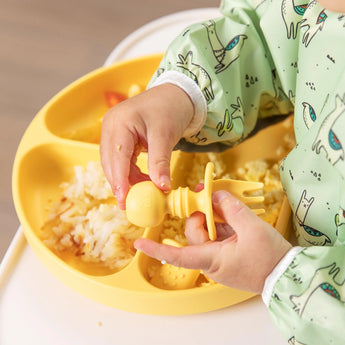 A child in dinosaur pajamas enjoys baby-led weaning, using Bumkins Silicone Chewtensils®: Pineapple to eat from a yellow divided plate.