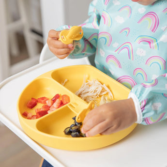 A child in a rainbow outfit uses Bumkins Silicone Chewtensils®: Pineapple at a high chair, eating from a silicone divided plate.