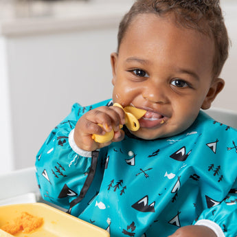 A baby in a highchair smiles with a Bumkins Silicone Chewtensils®: Pineapple spoon near their mouth, wearing a blue patterned bib.