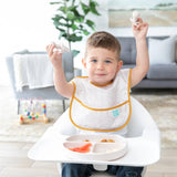 A toddler in a high chair holds Bumkins Silicone Chewtensils®: Sand, enjoying baby-led weaning, with toys scattered on the floor.