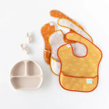 The Bumkins baby feeding set includes orange/yellow bibs, a beige divided plate, and Silicone Chewtensils®: Sand on white background.