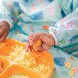 A child in colorful clothes enjoys scrambled eggs from a yellow divided plate using Bumkins Tangerine Silicone Chewtensils via baby-led weaning.