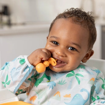 A smiling baby in a high chair, with a dinosaur bib, holds Bumkins Silicone Chewtensils®: Tangerine near their mouth.