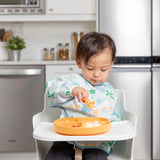 A toddler in a high chair wears a colorful bib, eating from an orange plate with Bumkins Silicone Chewtensils®: Tangerine.