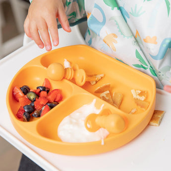 A child in a dinosaur outfit enjoys fruit, yogurt, & pancakes from an orange plate using Bumkins Silicone Chewtensils®: Tangerine.