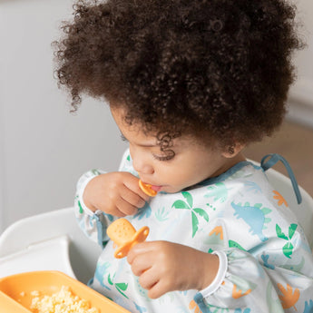 A curly-haired toddler enjoys a meal in a dinosaur bib using Bumkins Silicone Chewtensils®: Tangerine, sitting in a highchair.