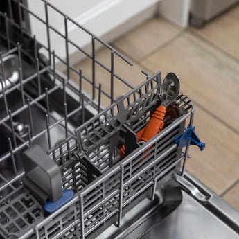 A close-up of Bumkins Spoon + Fork: Clay in a dishwashers basket showcases the orange-handled spoon, ideal for toddler self-feeding.