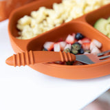 Orange divided plate with fruit, pasta, avocado. Bumkins Spoon + Fork: Clay set on the edge with silicone handle for self-feeding.