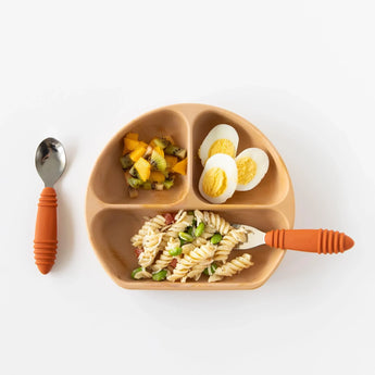 A toddler plate has sliced fruit, a halved egg, and veggie pasta. The Bumkins Spoon + Fork: Clay set is ready for self-feeding.