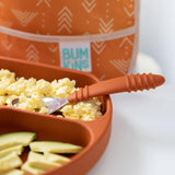 Close-up of a divided orange plate with scrambled eggs, avocado slices, and Spoon + Fork: Clay. A Bumkins bag is in the background.