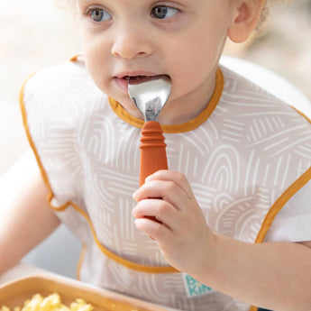 A toddler self-feeds scrambled eggs with Bumkins Spoon + Fork: Clay, using the orange-handled spoon while wearing a patterned bib.