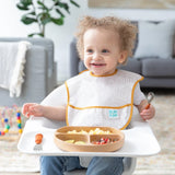 A toddler in a high chair uses their Bumkins Spoon + Fork: Clay set, holding a spoon with food on the plate and toys behind.