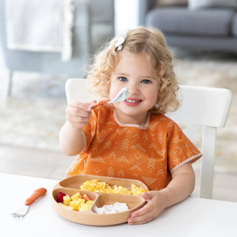 A curly-haired child eagerly self-feeds fruit and scrambled eggs with Bumkins’ Spoon + Fork: Clay, wearing an orange bib.