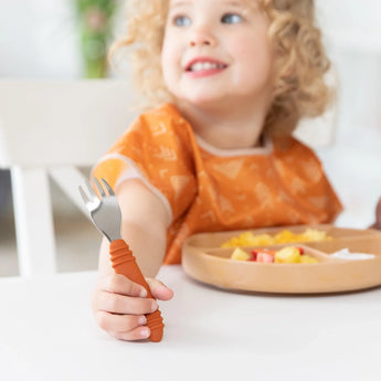 A smiling child in orange holds a spork from their Bumkins Spoon + Fork: Clay set, sitting at a table with diced fruit.