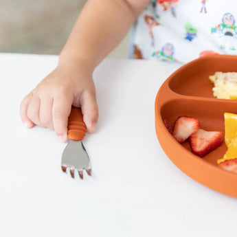 A childs hand reaches for strawberries, oranges, and eggs with a Bumkins Spoon + Fork: Clay utensil on a white table.