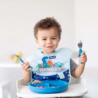 A toddler in a dinosaur bib labeled Hangry joyfully holds their Bumkins Spoon + Fork: Dark Blue over a plate of food.