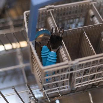 The Bumkins Spoon + Fork in Dark Blue and childrens utensils are in the cutlery basket of an open dishwasher.