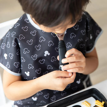 A child in a Mickey Mouse bib enjoys chocolate with Bumkins food-safe silicone Spoon + Fork, snacks on a tray nearby.