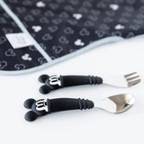 The Bumkins Mickey Mouse Spoon + Fork set with black cartoon designs rests on a patterned black mat.