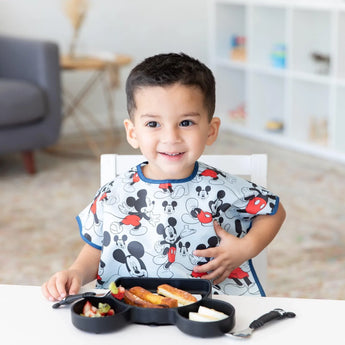 A young child smiles at the table wearing a Mickey Mouse bib with Bumkins Spoon + Fork: Mickey Mouse set and snacks in front.