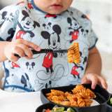 A child with a Mickey Mouse bib eagerly uses their Bumkins Spoon + Fork set to enjoy pasta from a divided plate.