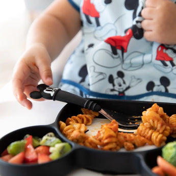 Child in Mickey Mouse shirt uses Bumkins Mickey Mouse silicone fork to enjoy pasta and veggies from a divided plate.