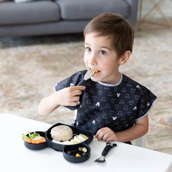 A child enjoys a meal with Bumkins Spoon + Fork: Mickey Mouse set, wearing a patterned bib and using a black lunchbox.