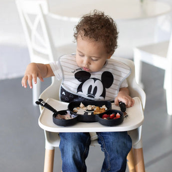 A toddler in a high chair, wearing a Mickey Mouse bib, enjoys snacks from their food-safe silicone divided plate with Bumkins utensils.