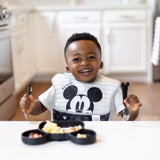 A smiling child uses Bumkins Mickey Mouse Spoon + Fork, eyeing the food-safe silicone plate shaped like Mickey Mouse.