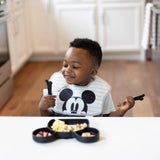 A child in a Mickey Mouse bib holds Bumkins Spoon + Fork: Mickey Mouse set, eager to dive into a Mickey-shaped meal.