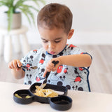 Toddler using Mickey Mouse Spoon + Fork by Bumkins eats pasta from a food-safe silicone plate, seated at the table.