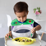 A toddler in a colorful bib uses Bumkins Jade Spoon + Fork to eat from a yellow plate in a high chair, with a nearby plant.