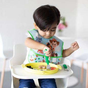 A toddler uses Jade Bumkins utensils with a cartoon bib, eating from a yellow plate.