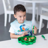 A young child in a bib smiles while using Bumkins Spoon + Fork: Jade to eat pasta and fruit from a green plate at the table.