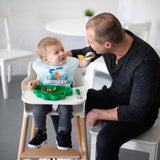 In a bright kitchen, a man feeds a smiling baby in a bib with Bumkins Spoon + Fork: Jade set as the baby reaches out.