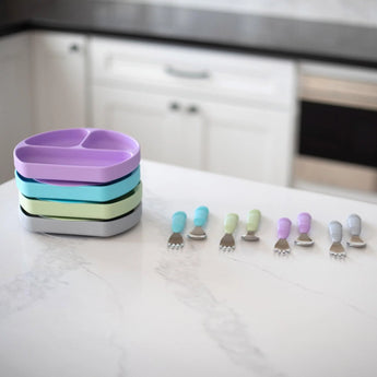 Lavender Spoon + Fork by Bumkins beside colorful kids plates on the counter with white cabinets behind.