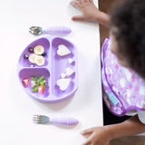 A child reaches for a lavender divided plate with fruits and sandwiches beside Bumkins Lavender Spoon + Fork self-feeding utensils.