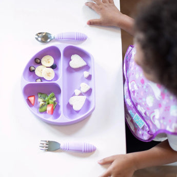 A child reaches for a lavender divided plate with fruits and sandwiches beside Bumkins Lavender Spoon + Fork self-feeding utensils.