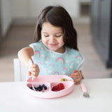 Young girl uses Bumkins Spoon + Fork: Pink to self-feed fruit from a divided pink plate with a rainbow-patterned bib at the table.