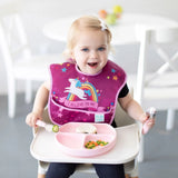 A toddler in a high chair, wearing a unicorn bib, eagerly self-feeding with Bumkins Spoon + Fork: Pink and a plate of food.