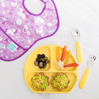 A yellow divided plate with toast, veggies, and blueberries is paired with a purple unicorn bib and Bumkins Spoon + Fork: Pineapple set.