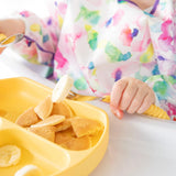 A child happily uses a Bumkins Spoon + Fork in Pineapple to enjoy pancakes and banana from a yellow divided plate.