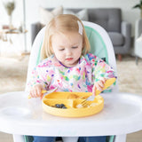 A toddler in a high chair uses the Bumkins Spoon + Fork: Pineapple set to enjoy blueberries and bananas from a colorful bib.